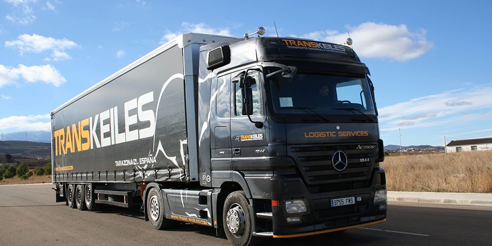 Tráiler de Transkeiles con lona negra rotulada, en un camino junto a un campo