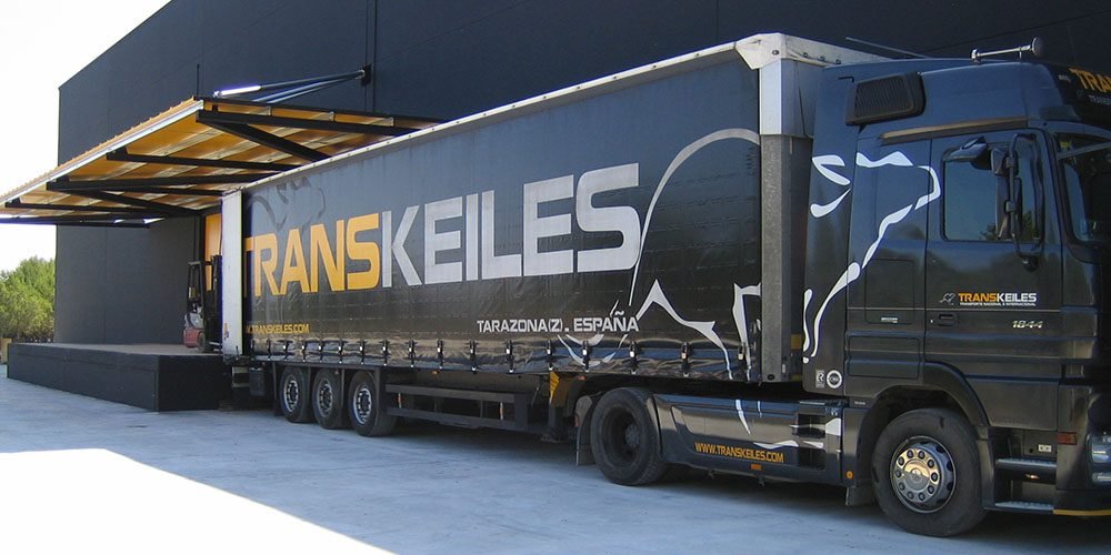 Tráiler negro de Transkeiles con la lona rotulada, aparcado en el muelle de carga de su nave